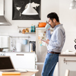Image of man wearing jeans and a button-up shirt listening to ear buds in white his kitchen while leaning against the counter drinking a cup of coffee.