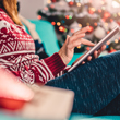 Image of woman wearing Christmas sweater and leggings, relaxing on the couch scrolling on a tablet in front of the colorful Christmas tree.
